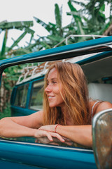 Red hair surf girl near blue vintage mini van with palm trees in the background in grey swimsuit, Indonesia, Bali © ArinaEmelyanova