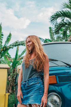 Red hair surf girl near blue vintage mini van with palm trees in the background in grey swimsuit, Indonesia, Bali