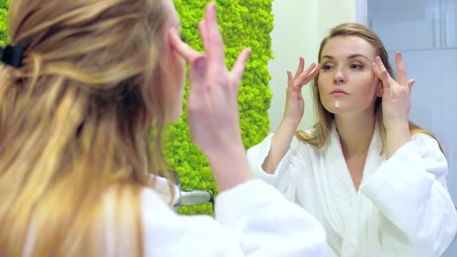 Pretty woman looking at mirror in modern bathroom and applying cream on face