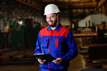 Man in a construction uniform, white construction helmet looking at the tablet and smiling at the background of the plant, production, industry