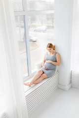 Young beautiful pregnant woman sitting on windowsill at home