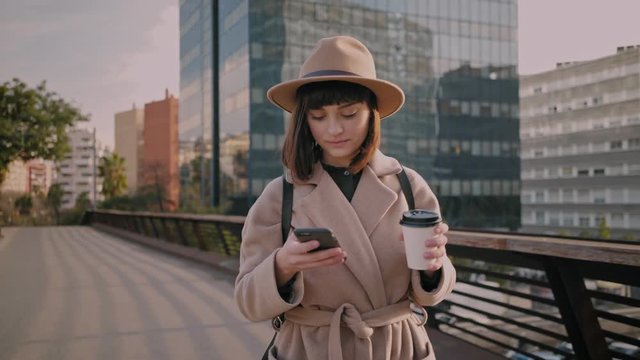 Attractive young businesswoman in fedora hat and fashionable coat walks on urban street and uses modern smartphone, slow motion