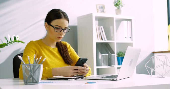 Attractive woman in glasses using smart phone while sitting at office desk in the business office. Close up business woman using cell phone indoors.