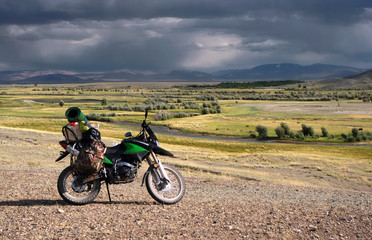 Motorcycle traveler enduro with suitcases standing on a stones in high mountain sunny dry desert steppe with yellow grass trees and bushes around river under the storm dark clouds Altai Siberia Russia
