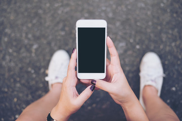 Mockup image of a woman sitting on the street and holding white mobile phone with blank white screen   