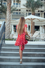 A girl with long straight hair in red dress with polka dots, bag and gold high-heeled shoes walking up the stairs