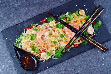 Noodles with prawns and vegetables on a black stone plate with traditional oriental sauce on a grey abstract background. Healthy food concept
