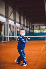  little boy on the yellow, sandy tennis court stands with racket in blue tracksuit