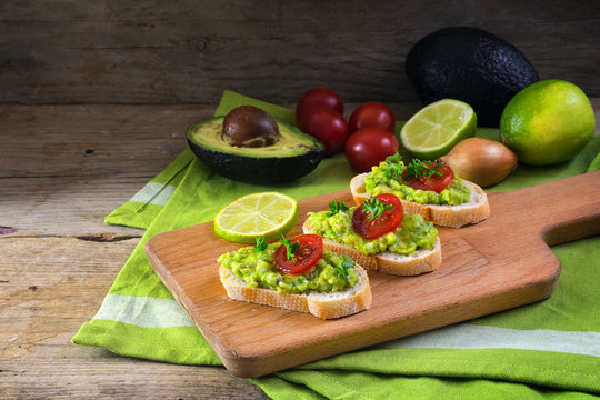 avocado cream or guacamole on baguette sandwiches with ingredients like lemon, onion and tomatoes on a rustic wooden board, healthy vegetarian party snack, copy space