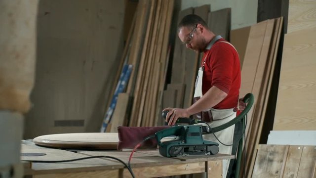 Carpenter with various electric sanders polishing a round plank in workshop