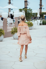 Beautiful young blond woman in a short translucent dress walking in the hotel at beach resort. Elegant luxury sexy blonde model wearing dress goes through the streets for shopping