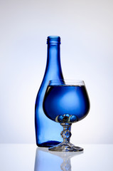 Transparent wine glass with water in front of a blue bottle on a white background