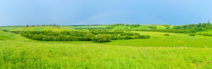Fototapeta na wymiar Wide panorama of hilly landscape with full rainbow in the sky, villages and church in background. 