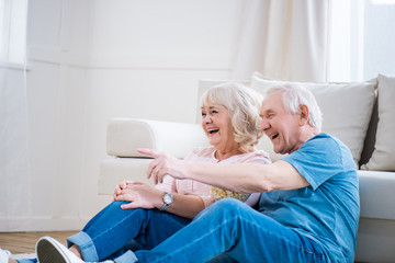 Happy senior couple laughing and sitting on floor, man pointing