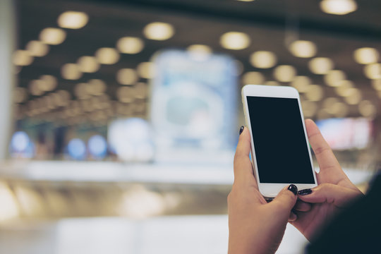 Mockup Image Of A Woman Holding And Using White Mobile Phone With Blank Black Screen While Standing And Waiting For Baggage Claim In The Airport