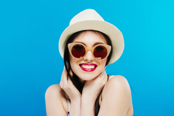 Portrait of Smiling Female Model in Fashion Sunglasses and Summer Hat on Blue Background.