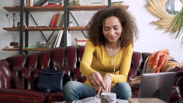 young latin curly woman speak with friend by telephone mobile using handsfree earpieces and look through magazine simultaneously while sitting and resting at home with cup of tea during sunny day
