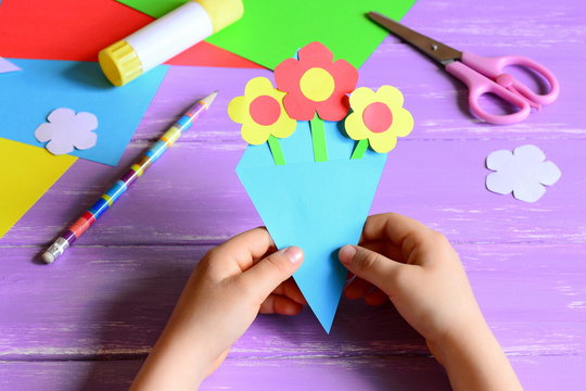 Little Child Made Paper Crafts For Mother's Day Or Birthday. Child Holds A Paper Bouquet In Hands. Easy And Beautiful Gift For Mom. Scissors, Glue Stick, Flowers Templates, Pencil On A Wooden Table
