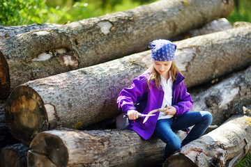 Cute little girl sitting on tree logs using a pocket knife to whittle a hiking stick - obrazy, fototapety, plakaty