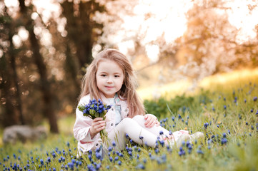 Smiling kid girl 4-5 year old holding blue flowers outdoors. Sitting in park wearing spring...