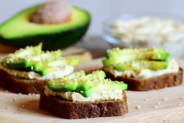 Healthy chickpeas hummus and avocado sandwiches on a wooden board, avocado half, hummus in a glass bowl. Veggie sandwiches made with rye bread, avocado slices, chickpeas hummus and fried sesame seeds