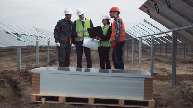 The group of engineers and workers in uniform on an incomplete field of photovoltaic panels discusses the plan of construction and mounting. They using blueprints and notebook