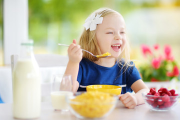Cute little girl enjoying her breakfast at home. Pretty child eating corn flakes and raspberries and drinking milk before school.