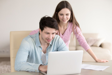 Cheerful young couple using laptop, man and woman discussing mortgage, investment planning, calculating and paying bills, analyzing their finances with documents to save money, working with computer