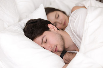 Young guy and his girlfriend sleeping together at home, man lying on pillow with smartwatch on his wrist, fitness tracker for easy awakening and healthy sleep, loving couple in comfortable soft bed
