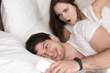Obraz na płótnie Canvas Surprised woman looking at her boyfriends mobile phone, when he texting or reading messages lying with his girlfriend in bed. Wife caught husband watching pornographic videos on smartphone