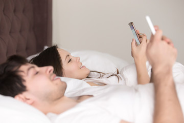 Obraz na płótnie Canvas Smiling young man and woman relaxing in comfortable bed holding cellphones using devices for online chatting, browsing websites, checking e-mail, blogging, mobile banking of shopping apps