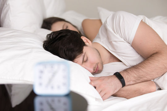 Young guy sleeping with girlfriend on cozy white bed, wearing smart watch that is better than alarm clock for healthy sleep, tracking and controlling awaking in the morning