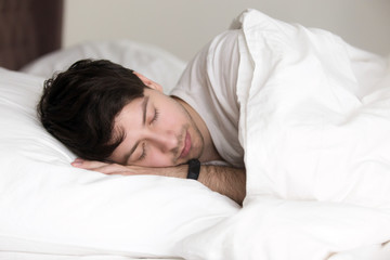 Fototapeta na wymiar Young handsome guy sleeping in soft bed under blanket, man asleep wearing activity tracker with sleep monitoring features on his wrist to feel full of energy after waking up