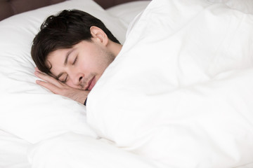 Fototapeta na wymiar Young guy sleeping alone in comfortable big bed at home or in hotel, enjoying white soft linen and downy bed, having a nap with eyes closed, pleasant morning for handsome single man