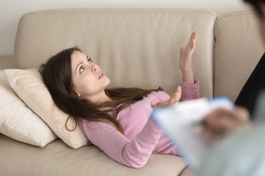 Beautiful young woman lying on couch at psychologists office, speaking about personal problems while doctor is making notes with clipboard and pen in hands, visit to professional psychotherapist