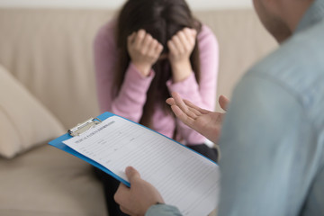 Young lady having depression sitting on the couch opposite to psychotherapist and crying. Male doctor holding clipboard intending to fill in all required information about medical patient