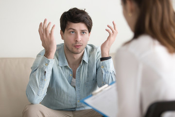 Man sitting on the couch at the psychologist, emotionally speaking, discussing with psychotherapist...