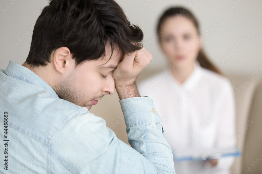 Wall mural Young worried guy touching his forehead, having a headache or migraine, overworked businessman feeling tired, stressed and exhausted, female doctor, nurse or colleague standing in the background