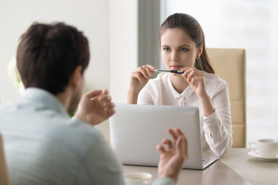 Young businesswoman looking serious and thoughtful, holding a pen near her mouth, listening attentively to self introduction of a male candidate during job interview or considering business offer