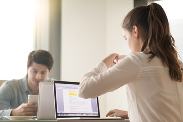 Young businesswoman working on laptop and businessman using smartphone sitting at the desk opposite her, colleagues drinking coffee, having a break or business lunch at the workplace in office