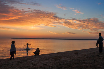Fototapeta premium Beautiful colorful summer sea sunrise landscape with amazing colorful clouds in a blue sky and unrecognizable people silhouettes waiting for the Sun. One man is splashing and jumping in water. In