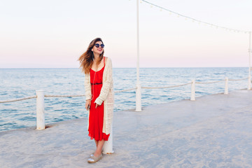 Beautiful girl in red dress and white jacket stands on a pier, smiles, and listens to music on...