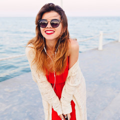 Close-up portrait of a beautiful girl in red dress and white jacket on a pier, smiling and listening to music on earphones on a smartphone. She wears dark sunglasses.