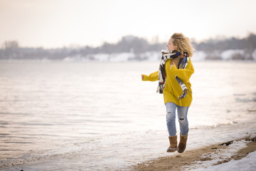 Beautiful blond woman in a yellow sweater and blue jeans running on the river shore. Happy girl...
