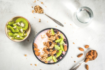 Healthy diet breakfast: smoothies bowl, with yogurt, fresh blueberries (black currant), kiwi, granola oatmeal, almond nuts. On a white stone table, with water and ingredients. Top view copy space