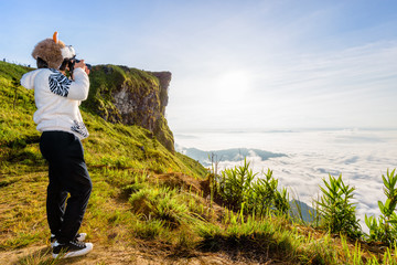 Asian hiker woman with digital camera photographing beautiful natural landscape of sky clouds and fog during sunrise in winter on mountain at Phu Chi Fa Forest Park, Chiang Rai Province, Thailand