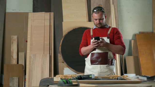 Carpenter wearing a woodworking apron typing a text message in a workshop