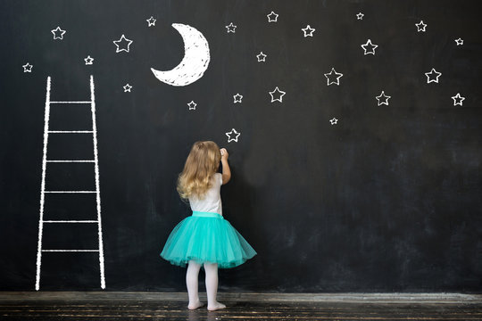 Cute little girl draws the moon and stars on the blackboard