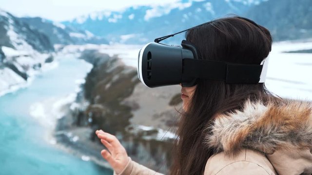 Young girl or teenager uses virtual reality glasses. Solves a puzzle or plays a game. Perhaps creating a design. Enjoys adventure and travel. On a background of winter mountains and a green river