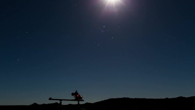 A static behind the scenes timelapse at night of a camera flashing on a linear dolly while shooting a landscape scene, with the moon and stars filing the sky
